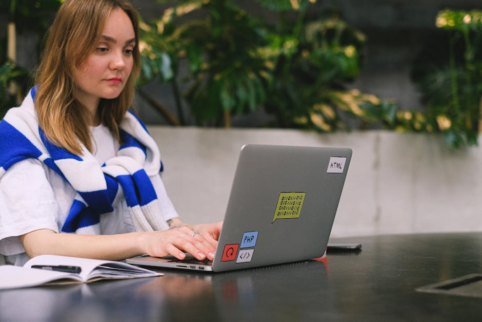 A young woman with shoulder-length blonde hair sitting at an outdoor table surrounded by lush green foliage, working on a silver laptop decorated with stickers labeled 'HTML', 'PHP', and others. She i